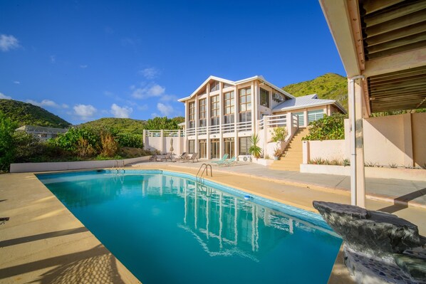 Une maison moderne dotée d’une piscine, d’un escalier menant à un balcon et offrant une vue sur une colline verdoyante.