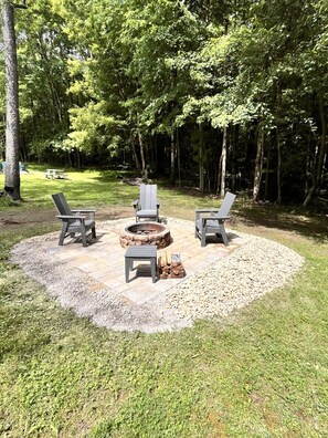 Un espacio de estar al aire libre con forma circular, que incluye un brasero rodeado por un anillo de grava y tres sillas Adirondack.