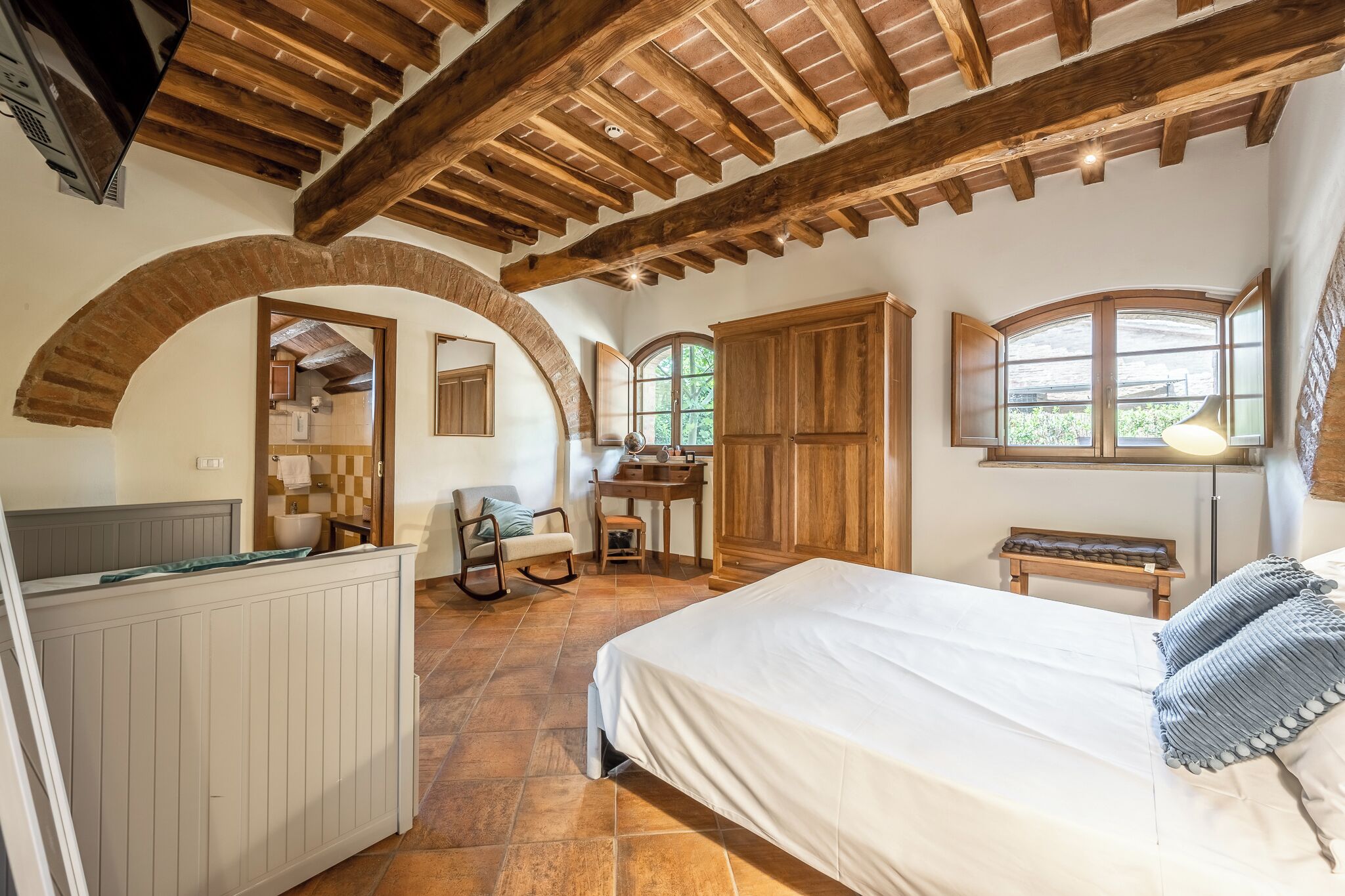 A bedroom with a large bed, wooden ceiling beams, a desk, and a chair.