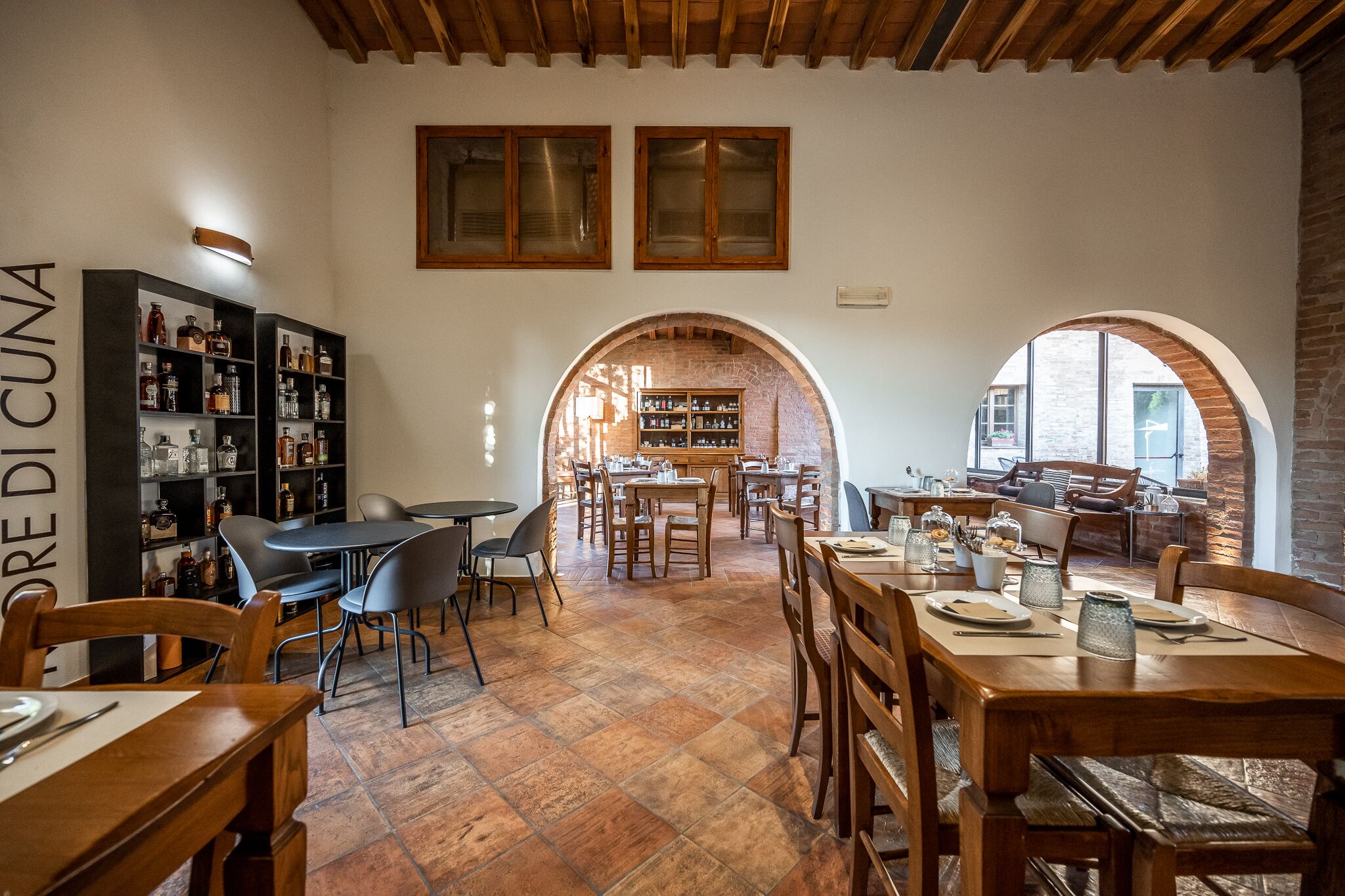 Un restaurant acollidor amb taules i cadires de fusta, una paret de maó i prestatges plens de botelles.
