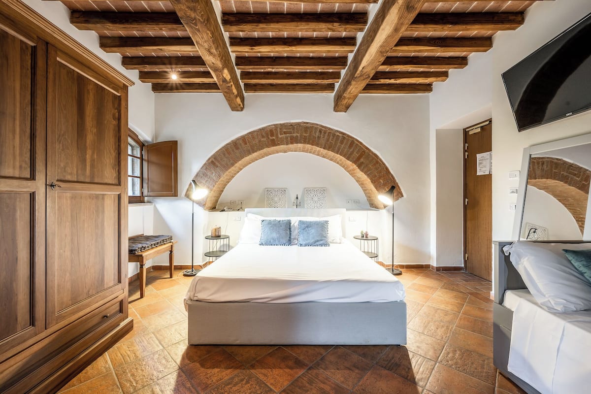 Un dormitorio con una cama grande, vigas de techo de madera, techo de ladrillo abovedado y un televisor en la pared.