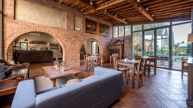 A spacious indoor area with a brick archway leading to a kitchen, wooden tables and chairs, and large windows offering a view of the outdoors. - Hotel More Di Cuna (Monteroni d'Arbia)