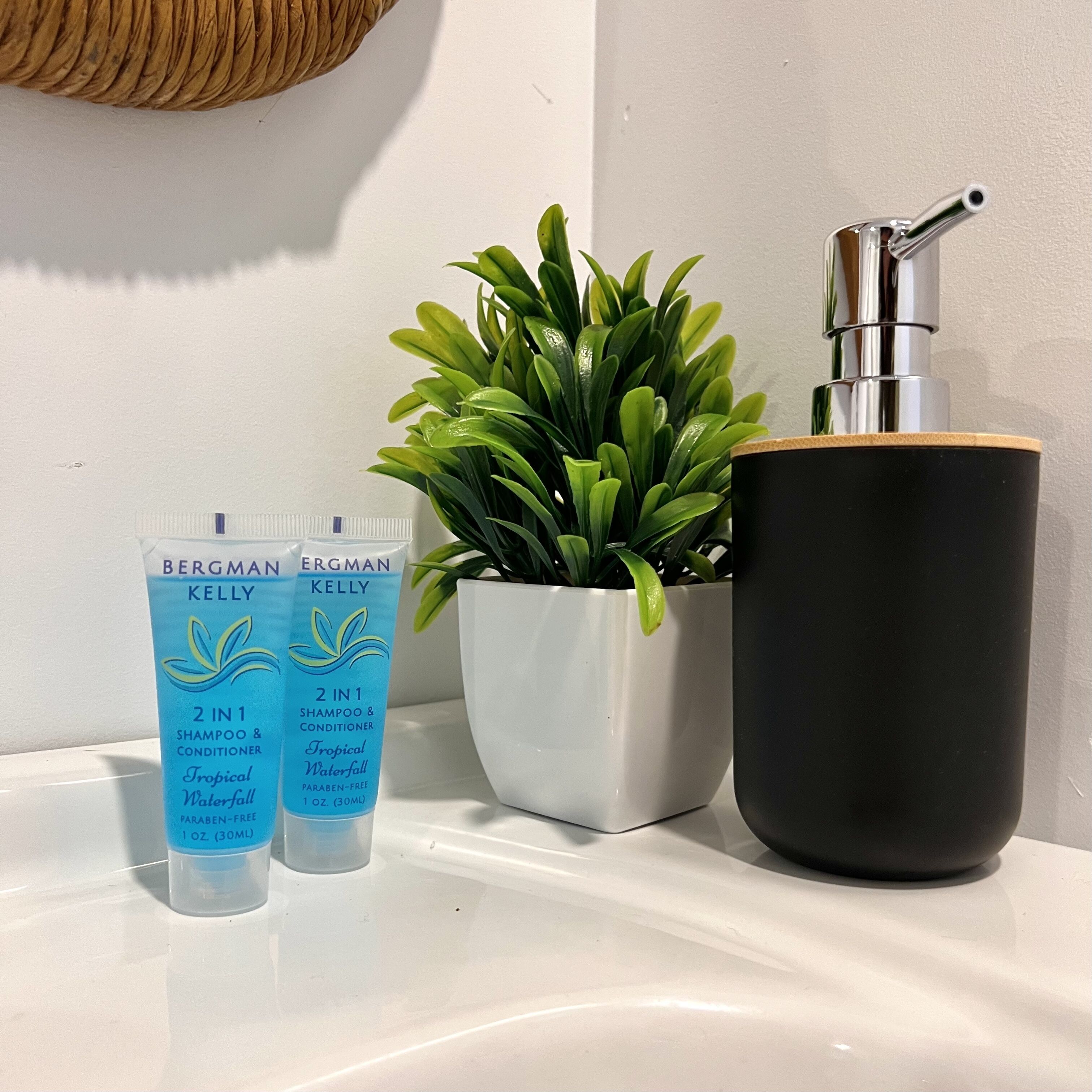 Kylpyhuoneen lavuaarin päällä on kaksi Bergman Kelly -merkkistä 2-in-1-shampoo- ja hoitoainepulloa, ruukkukasvi ja musta saippua-annostelija.