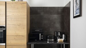 Une cuisine compacte équipée d’un four à micro-ondes, d’un grille-pain et d’une machine à café, le tout posé sur un plan de travail noir. Les meubles sont en bois et un tableau encadré orne le mur.