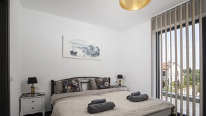 Une chambre à coucher avec un lit, deux tables de chevet, une lampe, un tableau et une vue dégagée sur l’extérieur grâce à de grandes fenêtres.