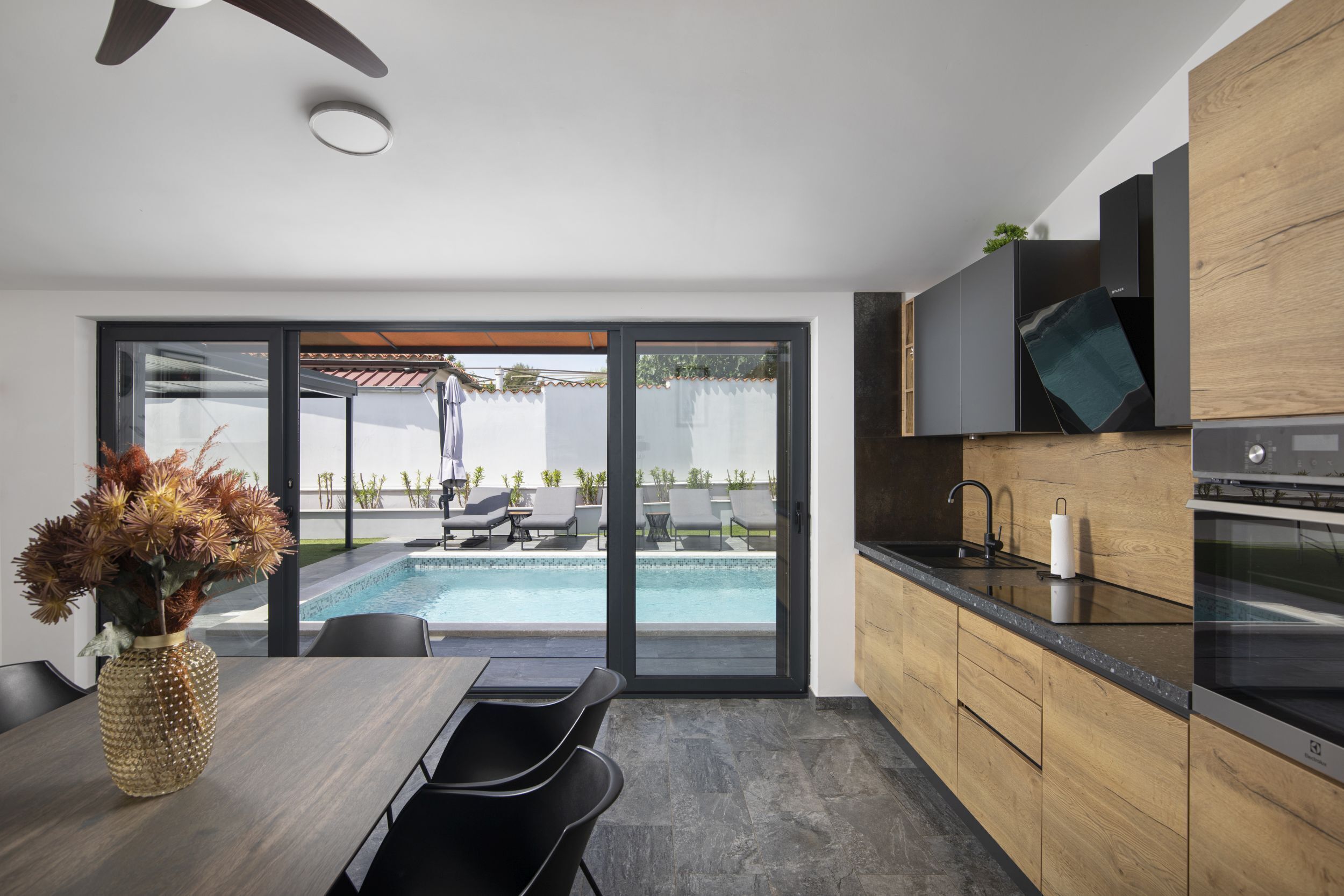A modern kitchen with a built-in oven, a dining area with a table and chairs, and a view of a swimming pool and outdoor seating.