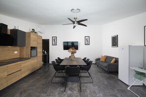 A modern kitchen with a dining area, featuring a central table, black chairs, a grey sofa, and a ceiling fan.