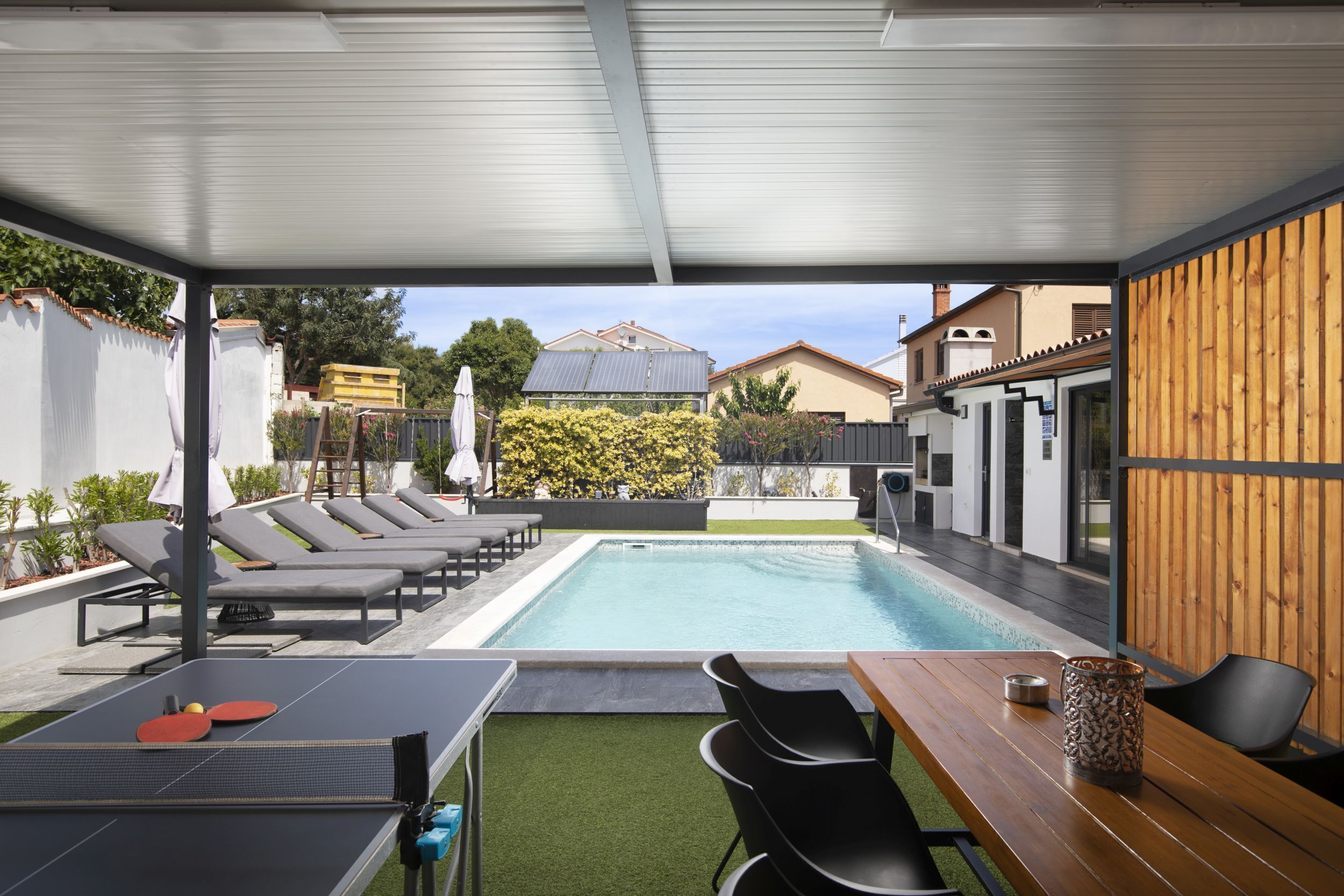 An outdoor pool area with lounge chairs, a ping pong table, and a wooden table with chairs.