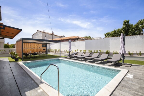 A swimming pool with lounge chairs and a table, surrounded by a white wall and a wooden fence.