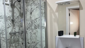 Una ducha con cerramiento de vidrio, una pared de mármol, un lavamanos blanco y un espejo sobre él.