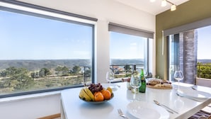 A dining area with a table set for two, featuring a fruit bowl, cheese, wine glasses, and a view of the countryside.