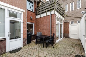Een bakstenen huis met een witte deur, een klein terras met een tafel en stoelen, en een houten omheining.