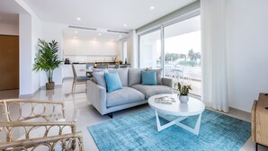 A modern living room with a sofa, coffee table, and wicker chairs. A kitchen is visible in the background with white cabinets and a dining area. - Adega Marina Apartments - Urban View (Lagos)