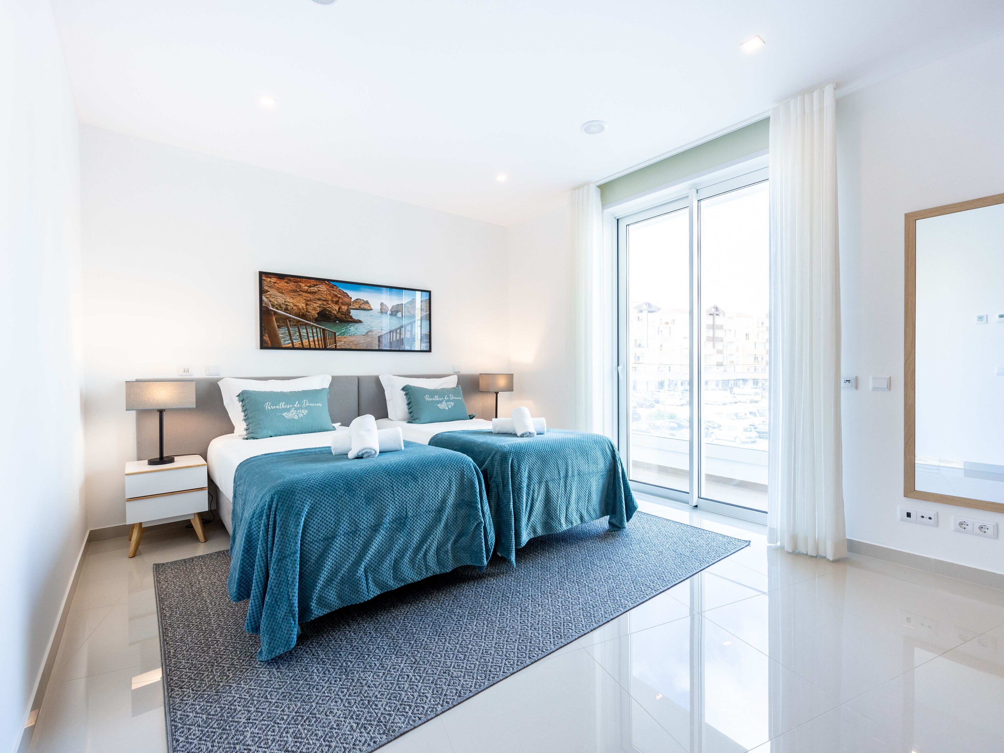 A modern hotel room with two beds, a large window, and a painting of a coastal scene.