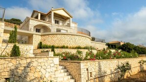 A stone-walled building with a curved balcony, a staircase leading up to it, and a garden with flowering plants. - Villa Gionis (Evreti)