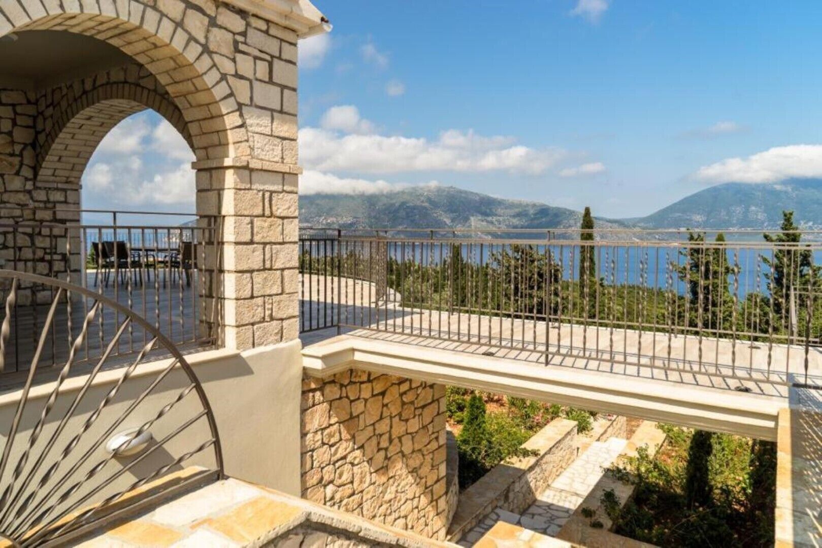 A stone balcony with a metal railing overlooking a mountainous landscape and a body of water.