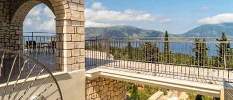 A stone balcony with a metal railing overlooking a mountainous landscape and a body of water.