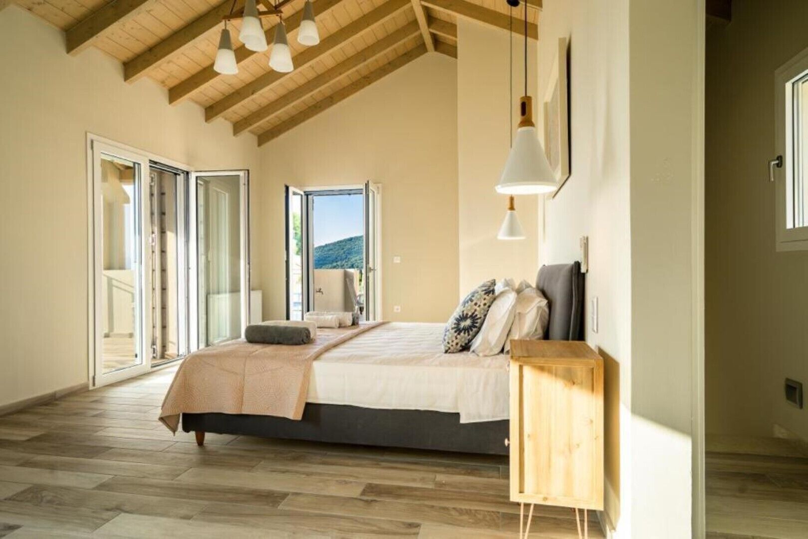 A bedroom with a large bed, wooden flooring, a wooden ceiling, and a view of the outdoors.