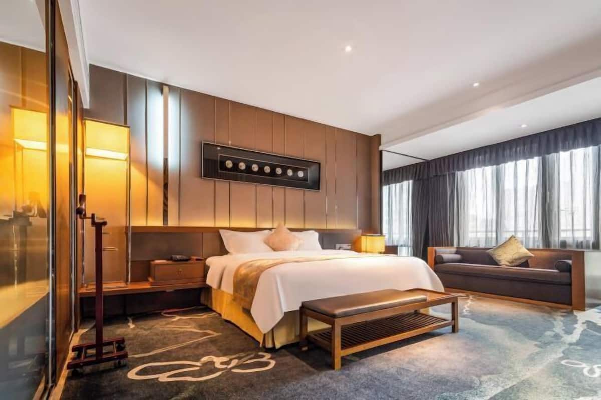 A modern hotel room with a large bed, a sofa, a desk, and a large window with curtains.