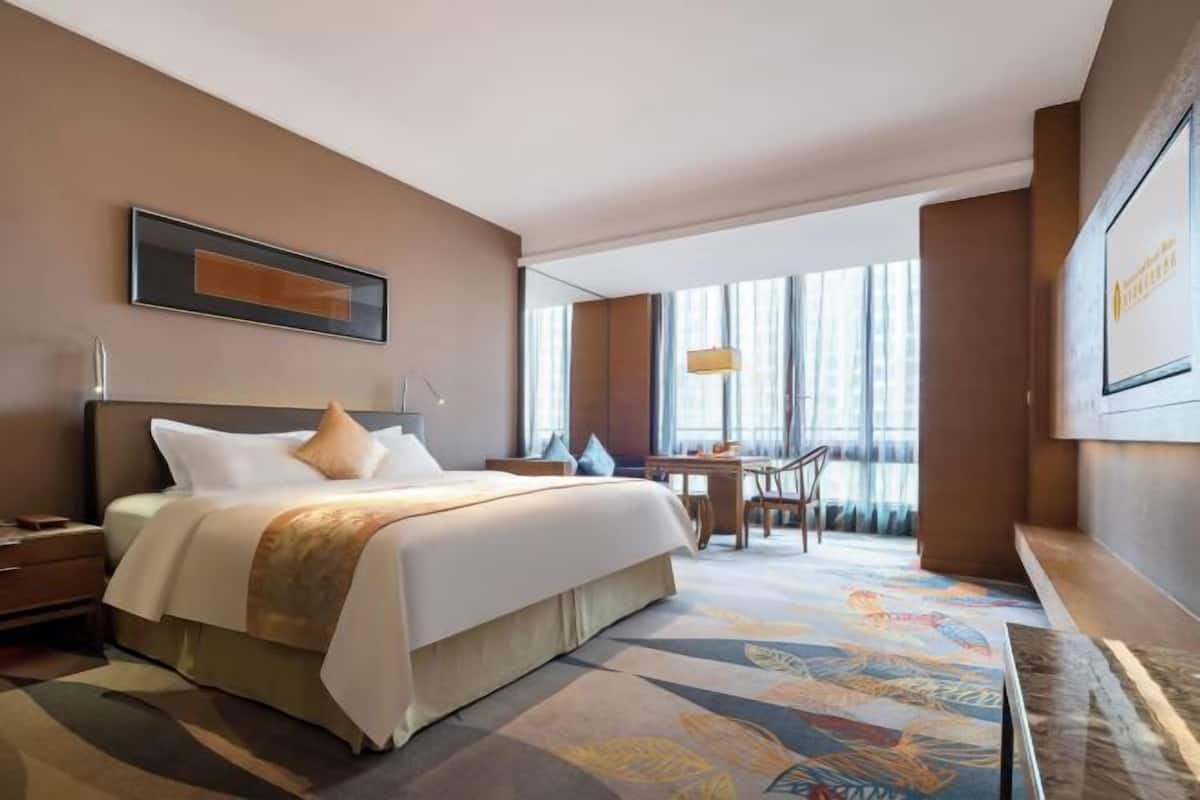 A hotel room with a bed, bedside table, desk, chair, and large windows.