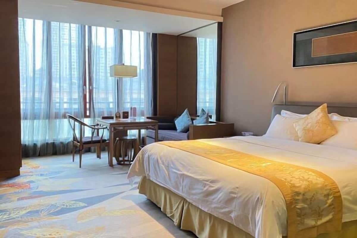 A hotel room with a large bed, a desk with chairs, a sofa, and a view of buildings through large windows.