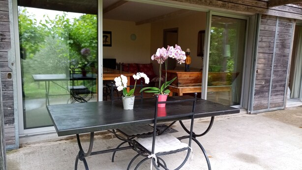 An outdoor dining area with a table and chairs, potted orchids, and a sliding glass door leading to an interior space. - 6/8 person gîte, all comforts in a family gîte village (Les Vans)