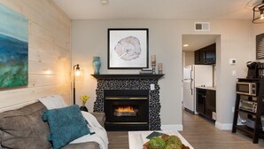 Una sala de estar con chimenea, un sofá, una mesa de centro con adornos y una cocina al fondo.