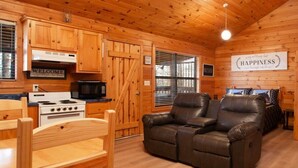 A cozy cabin interior with wooden walls and floors, a dining table with chairs, a sofa, a kitchen area with a stove and microwave, and a 'Happiness' sign on the wall. - Beautiful Mountain Cabin/Hot Tub (Sevierville)