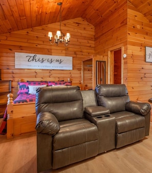 A cozy room with a leather sofa, wooden walls, and a chandelier. A sign on the wall reads "this is us." - Gorgeous Mountain Views/Hot Tub (Sevierville)