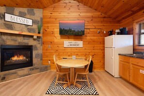 A cozy cabin interior with a stone fireplace, a dining table with chairs, a kitchen area, and a television mounted on the wall. - Gorgeous Mountain Views/Hot Tub (Sevierville)