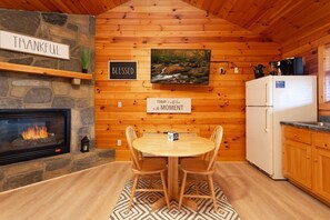 A cozy cabin interior with a stone fireplace, wooden walls, a dining table with chairs, and a refrigerator.