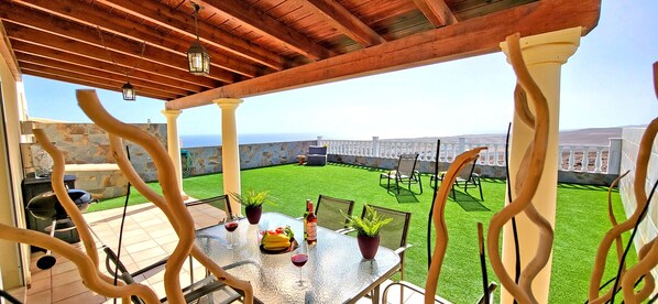 Een overdekt terras met een eettafel, een barbecue en uitzicht op een groen gazon en een witte schutting.