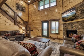 A log cabin interior with a fireplace, a staircase, and a large window.