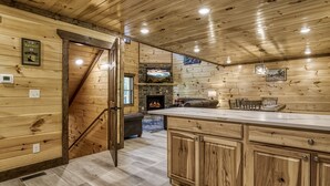 A log cabin interior with a stone fireplace, wooden staircase, and a kitchen area with wooden cabinets.