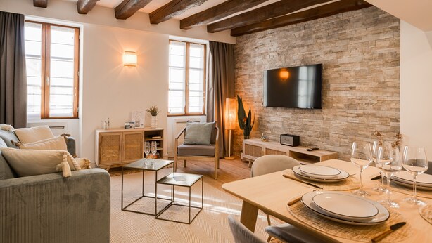 A modern living room with exposed wooden beams, a stone wall, a flat-screen TV, and a dining area set for six.