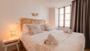 A neatly made bed with a white bedspread and pillows, a bedside lamp, and a window with curtains.