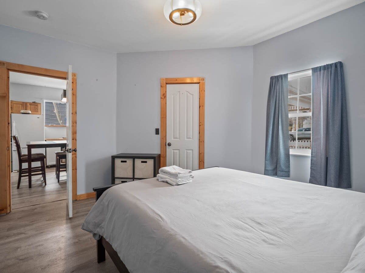 Ein Schlafzimmer mit einem Bett, einer Tür, einem Fenster mit Vorhängen, einem Schreibtisch mit Stühlen und Blick ins Freie.