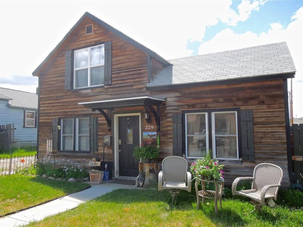 A two-story wooden house with a front porch, two chairs, and a small table with plants.