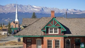 A building with a prominent steeple and a brick facade, set against a mountainous backdrop. - The Depot — Downtown Leadville Victorian, 3 Queens, Mt Elbert Views, Jetted Tub (Leadville)