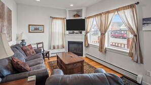 A living room with a sofa, a television, a fireplace, and large windows with curtains. - The Depot — Downtown Leadville Victorian, 3 Queens, Mt Elbert Views, Jetted Tub (Leadville)
