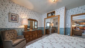 Un dormitorio con una cama grande, un aparador de madera y una silla. Hay un espejo sobre el aparador y un cuadro en la pared.