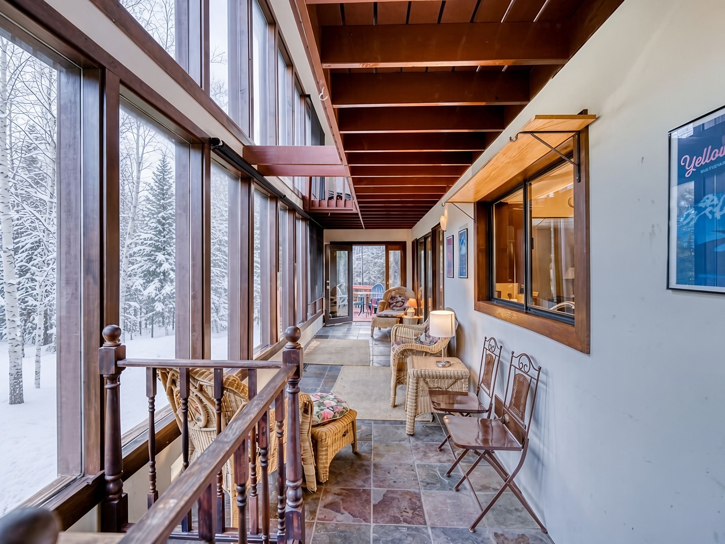 A covered outdoor area with a wooden staircase, wicker furniture, and large windows overlooking a snowy landscape.