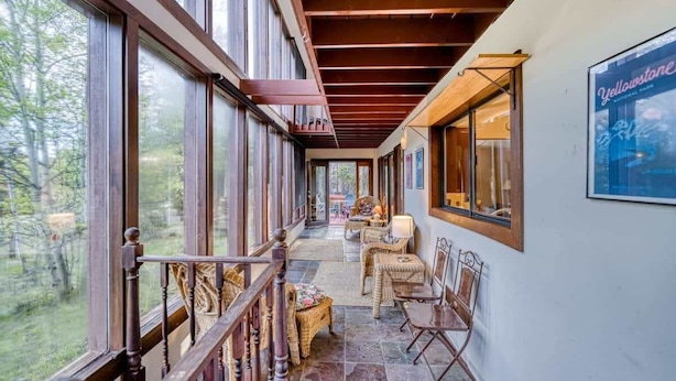 Un espacio al aire libre techado con barandilla de madera, mobiliario de mimbre y amplias ventanas que dan a un paisaje exterior.