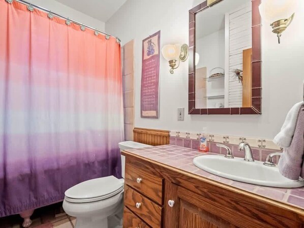 Un baño con cortina de ducha degradada, un inodoro blanco, un mueble de tocador de madera con un lavabo y un espejo sobre el mueble.