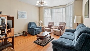 A living room with a wooden floor, a chandelier, a television, and a large window with blinds. - The Painted Lady — Victorian 4BR Steps to Main St Leadville, Sleeps 10, Dogs OK (Leadville)