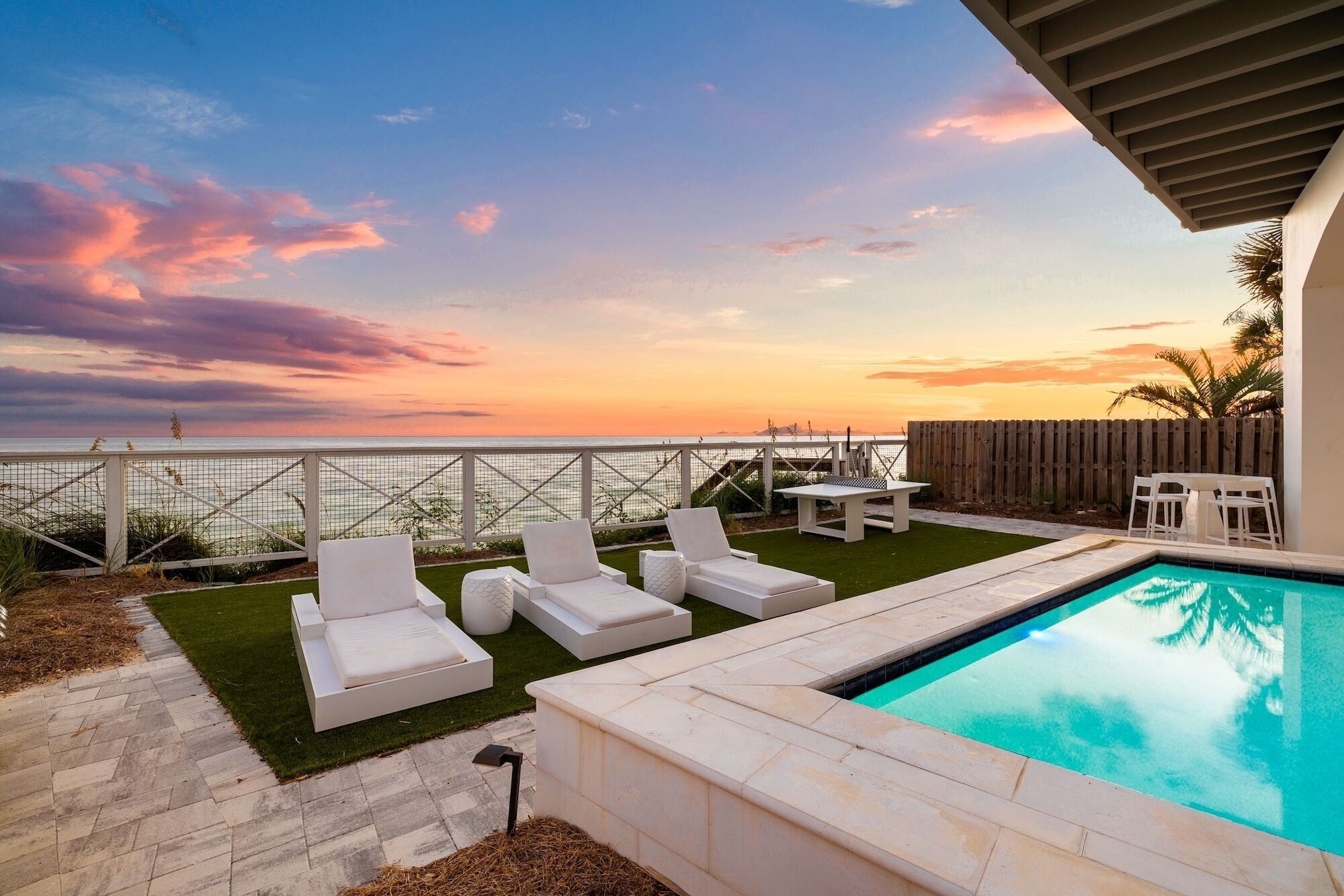 A pool area with lounge chairs, a small table, and a view of the ocean at sunset.