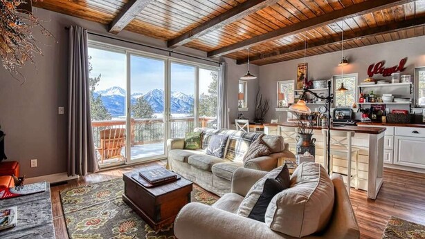 A spacious living area with a large window overlooking mountains, a cozy seating area with a fireplace, and a modern kitchen with white cabinets and wooden accents. - Mountain-View Cabin w/ Fireplace & Deck (Twin Lakes)