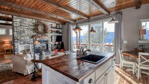 A rustic kitchen with a stone wall, wooden beams, a large island with a sink, and a dining area with a view. - Mountain-View Cabin w/ Fireplace & Deck (Twin Lakes)