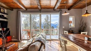 A cozy living room with a sofa, a wooden dining table, and a large window with a mountain view. - Mountain-View Cabin w/ Fireplace & Deck (Twin Lakes)
