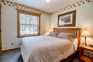 A bedroom with a bed, two bedside lamps, a framed picture on the wall, and a window with blinds.